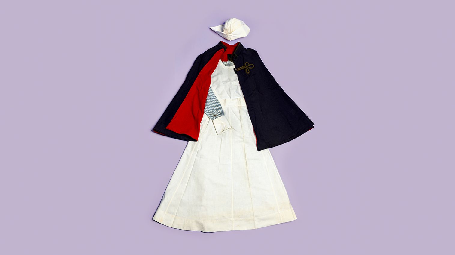 Nursing uniform consisting of a white hat, blue striped shirt, white apron, and navy cape with red lining, against a light purple background