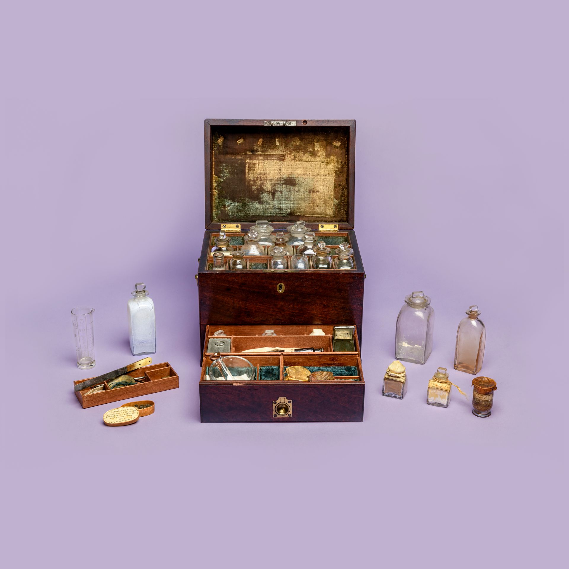 Brown wooden chest displayed open with its contents arranged next to it, including small glass vials and jars, against a light purple background